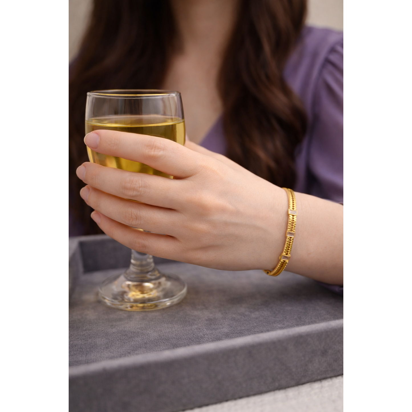 Gold lock bracelet with rectangular crystal accents worn on wrist holding small goblet with apple juice on grey velvet tray