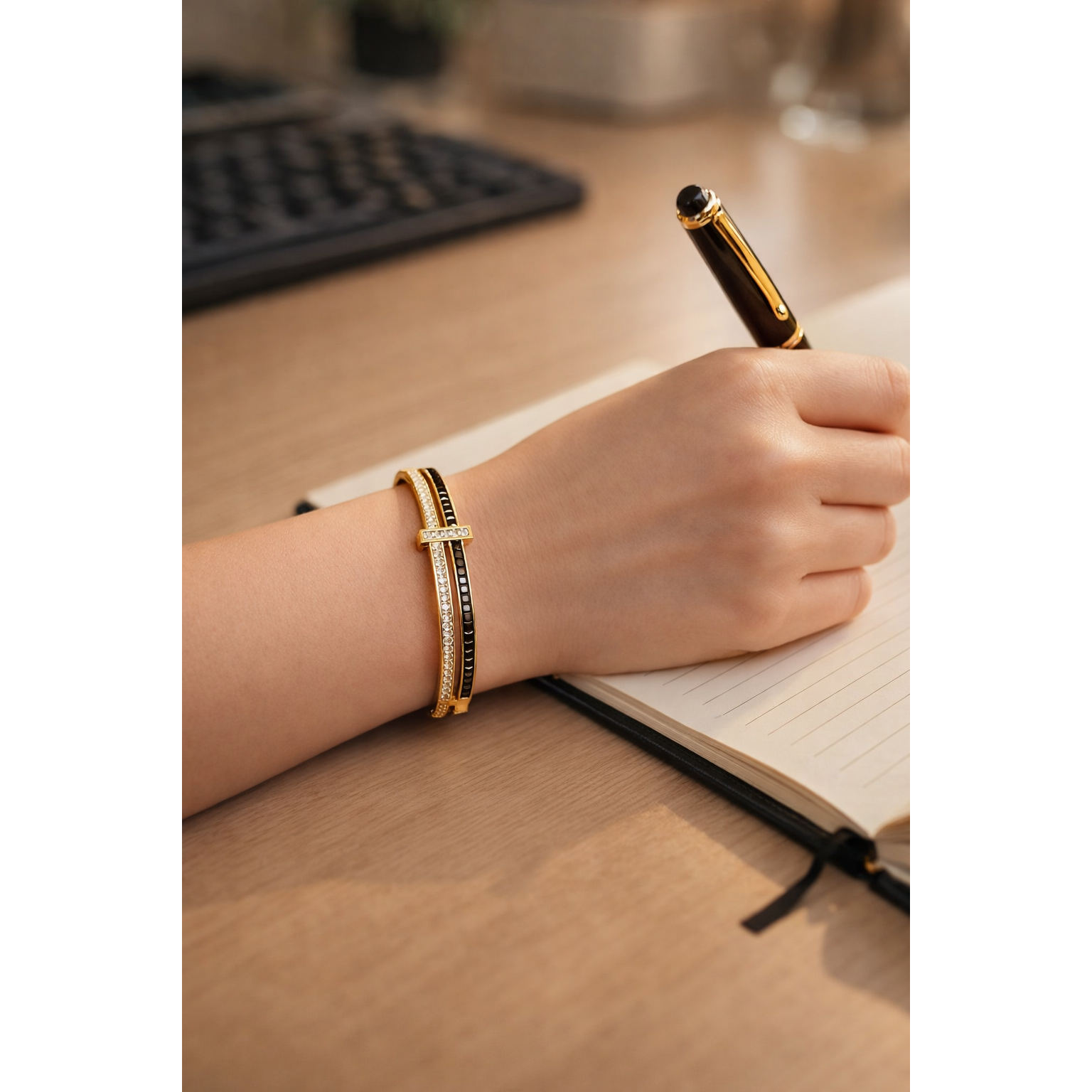 Gold lock bracelet with crystal and black stone detailing worn on wrist while writing in notebook on office desk