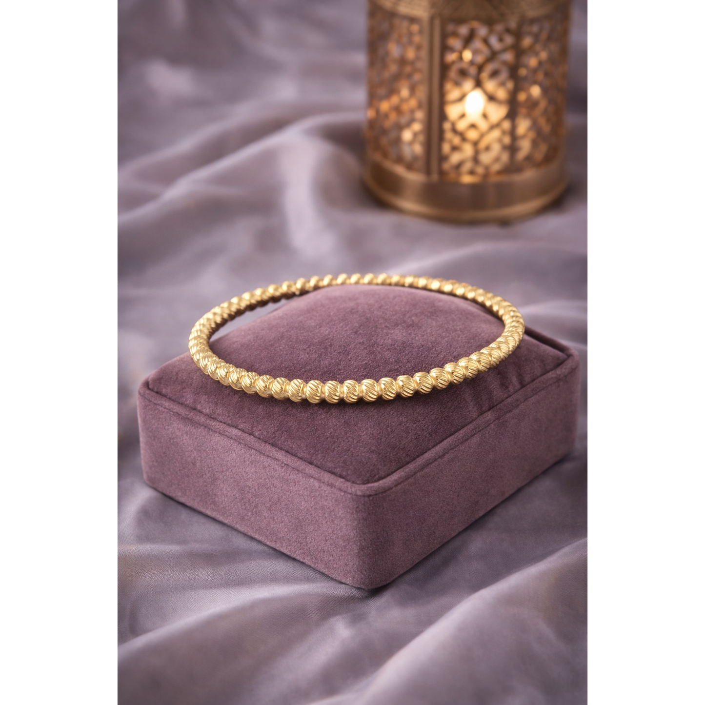 Gold textured bracelet with lock displayed on mauve velvet jewellery cushion with soft satin background and warm lantern ligh