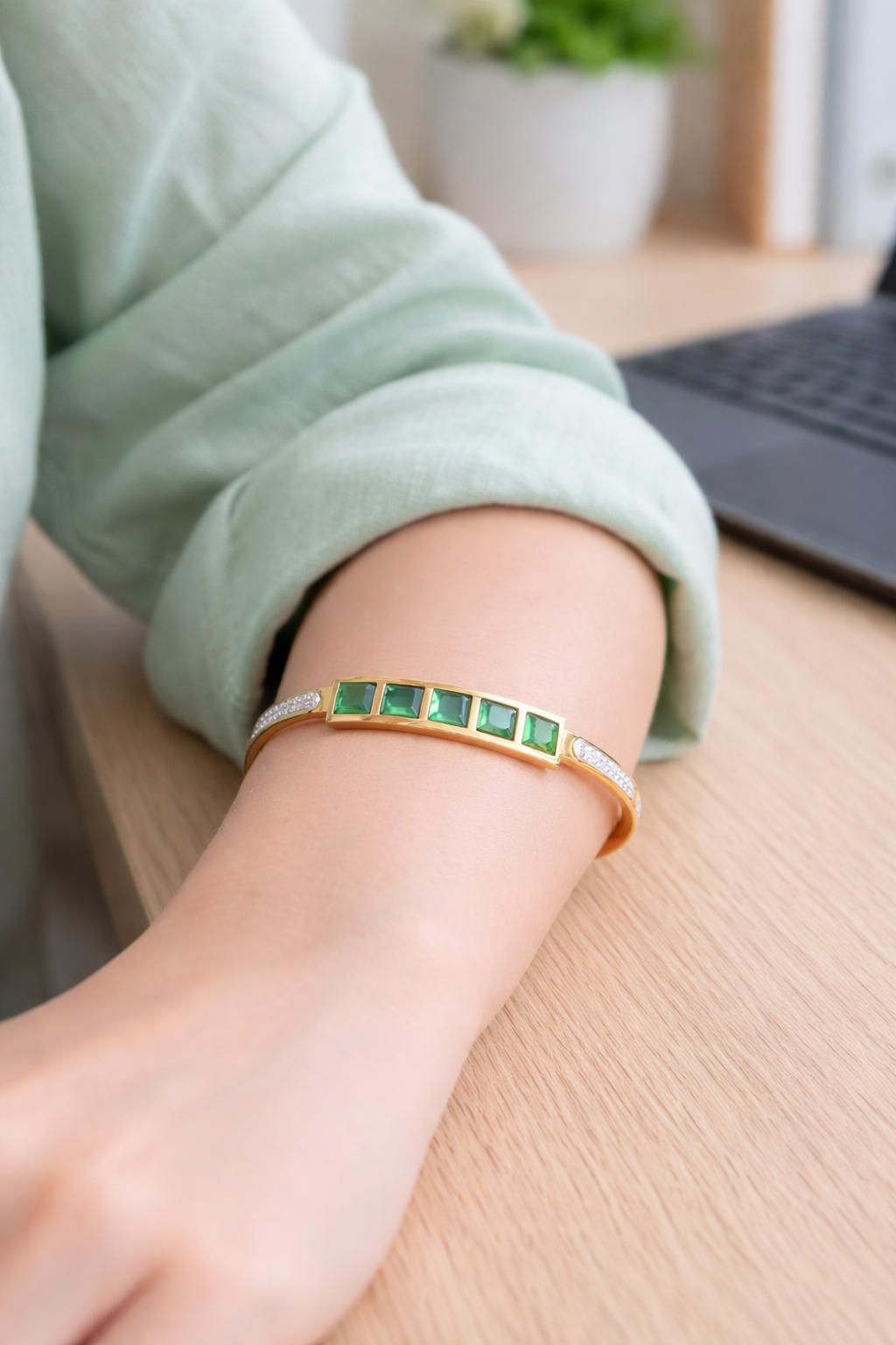 Gold bracelet with emerald green square stones and zircon accents worn on wrist on a work desk with blurred laptop background