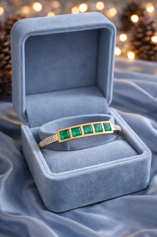 Gold bracelet with emerald green stones placed in a smokey blue velvet jewelry box on satin fabric with fairy lights and pine cones