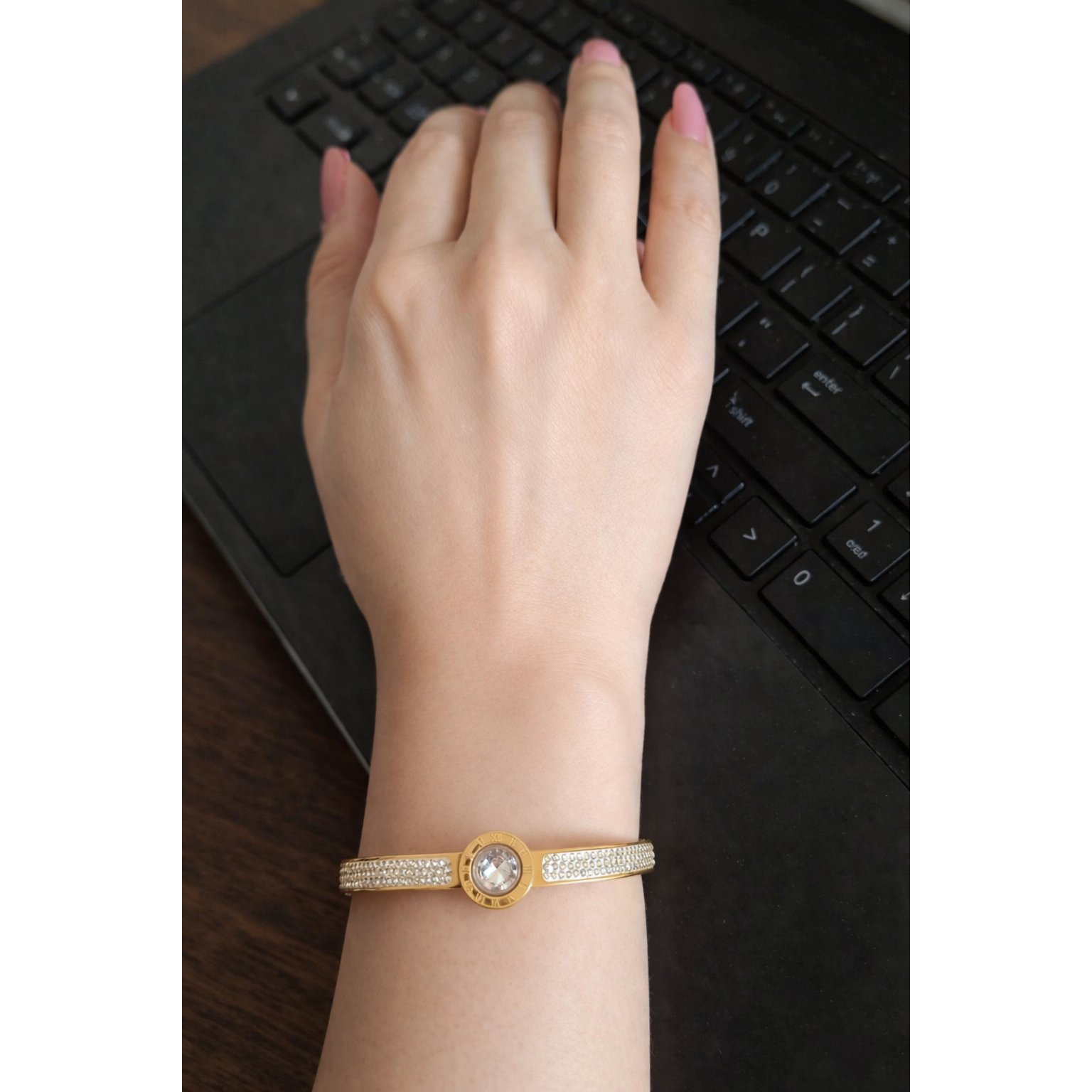 Gold Roman dial crystal bracelet with lock worn on feminine hand with pink nails on laptop workspace