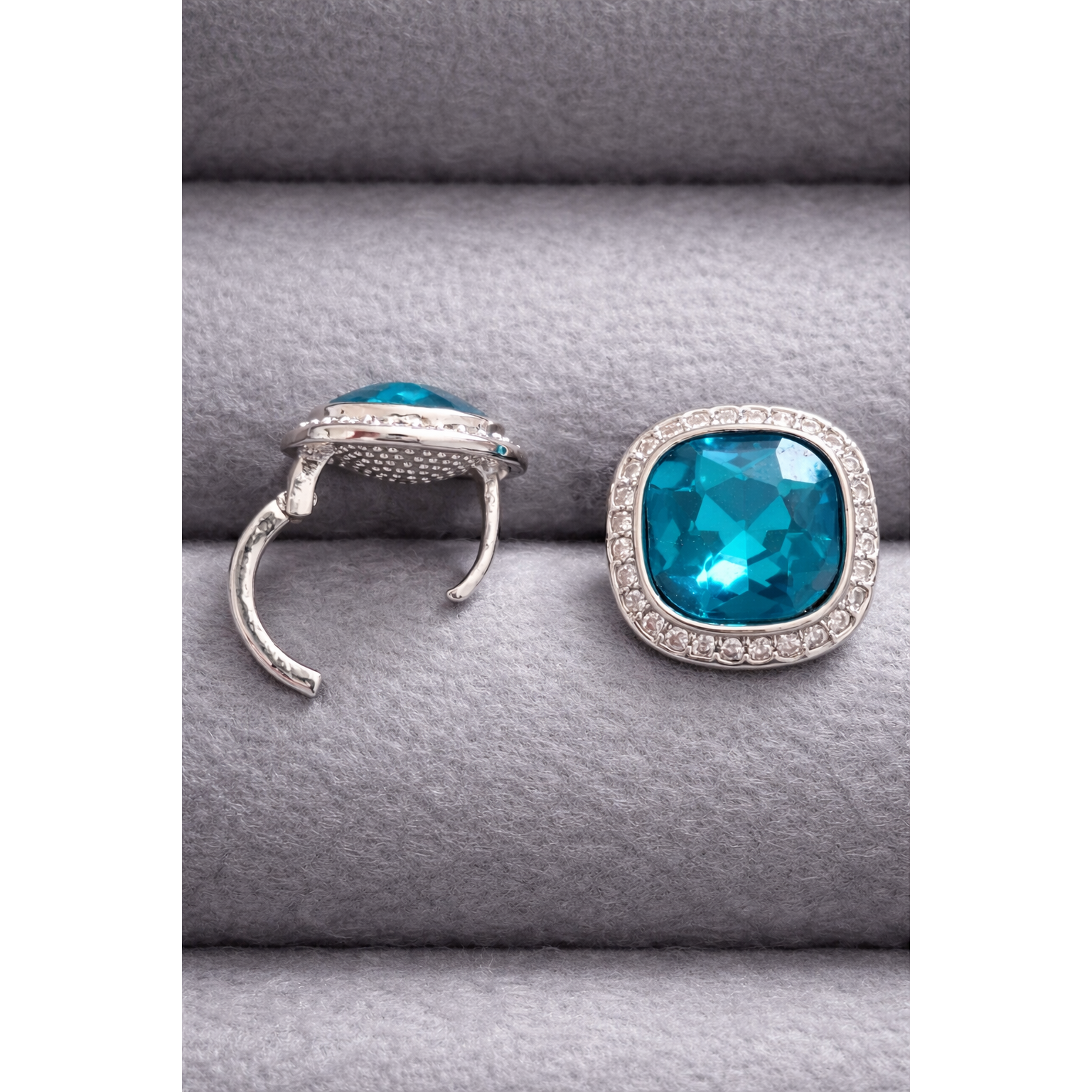Silver cushion-cut turquoise crystal stud earring shown in side and front position with halo stones on grey velvet display.