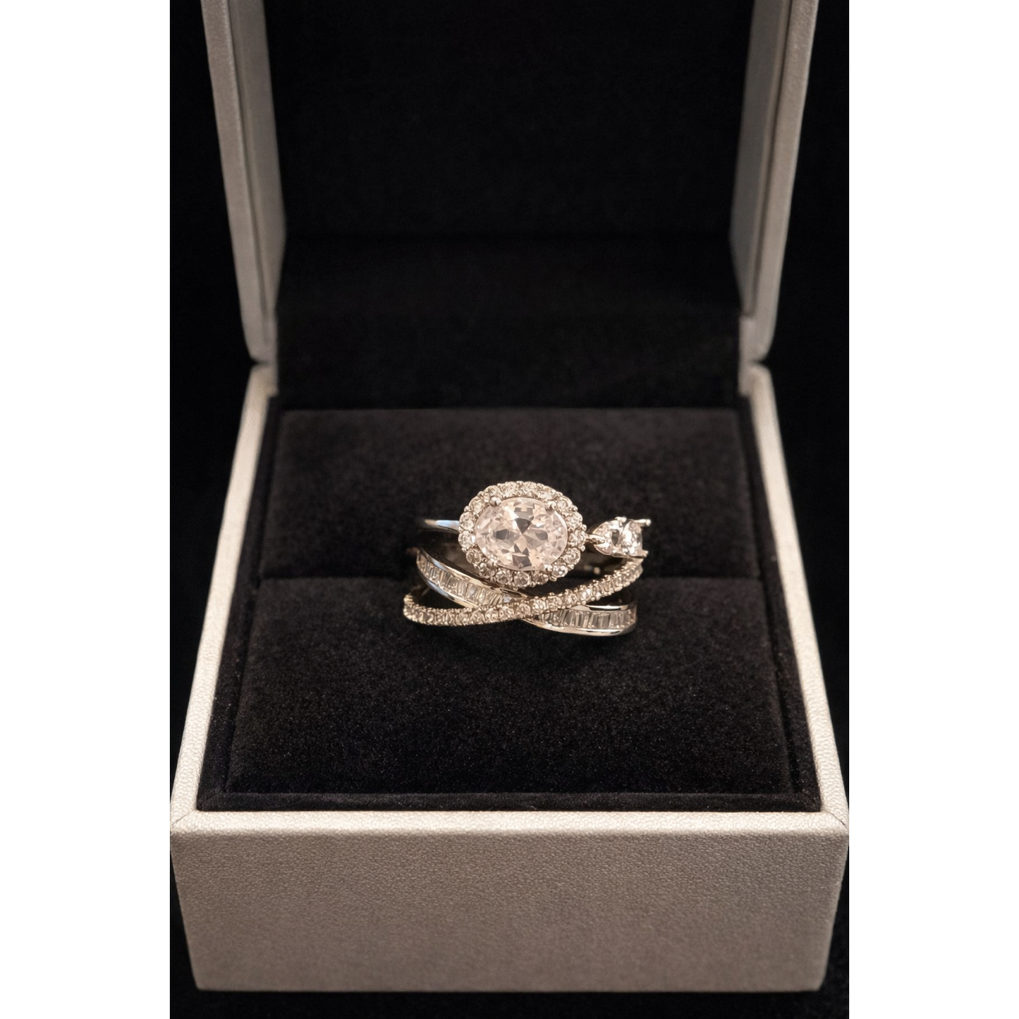Oval halo crystal ring displayed inside luxury jewelry box with black velvet interior, showing layered baguette-style and pavé stone band in silver metal