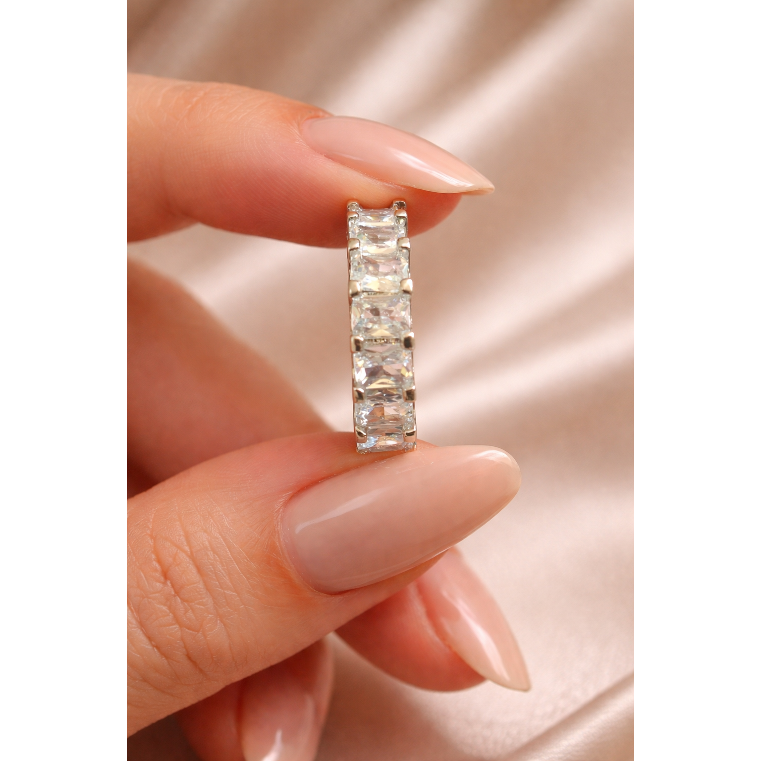 Silver baguette-cut crystal eternity band held between feminine fingers with beige nail polish, studio lighting, silk background