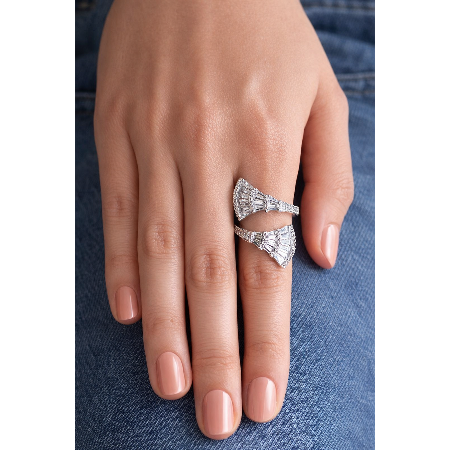 Silver tone open wrap fan-shaped baguette statement ring worn on female hand with peach pink nails against blue denim background