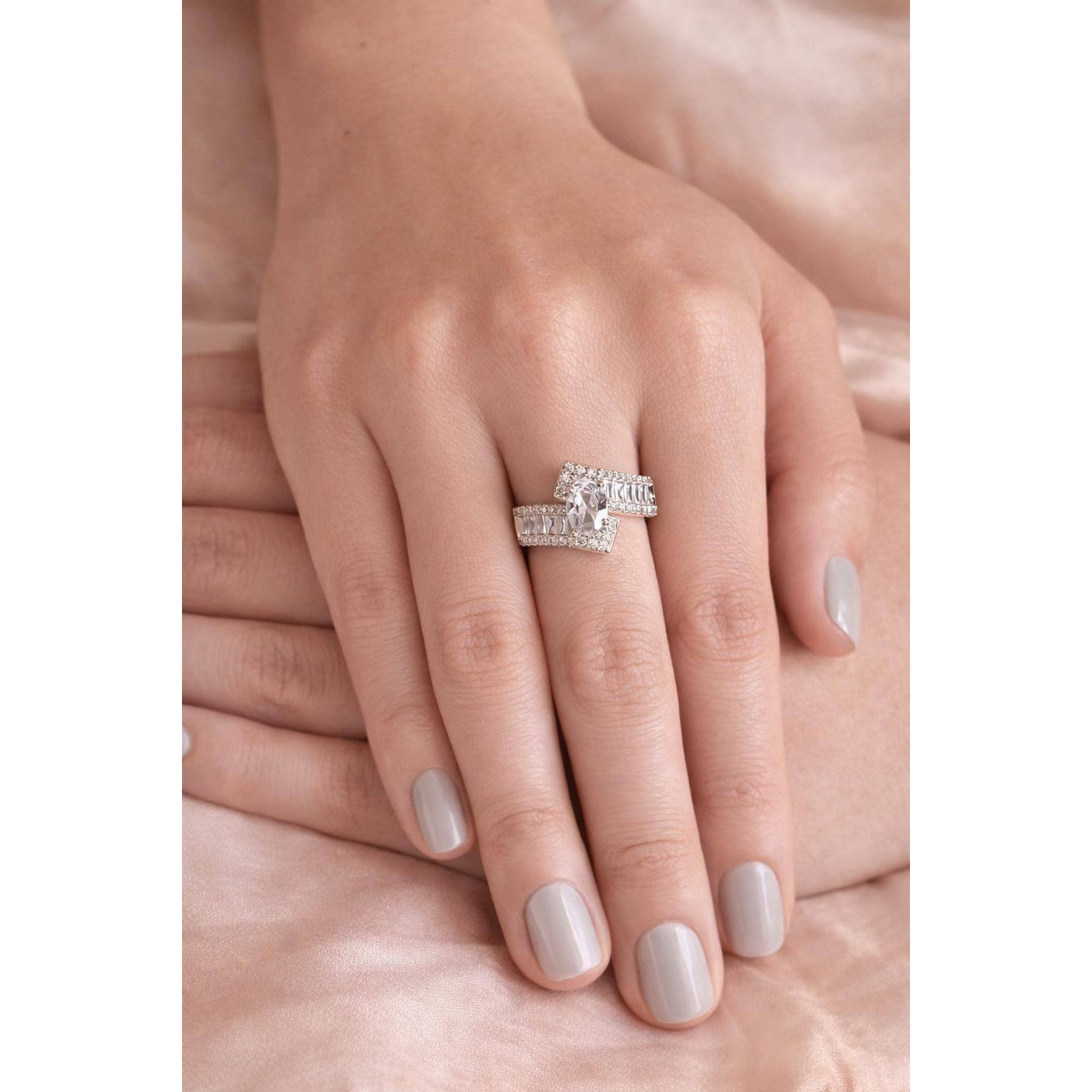 Silver oval center stone double-band statement ring worn on female hand with light grey nails on peach silk fabric background