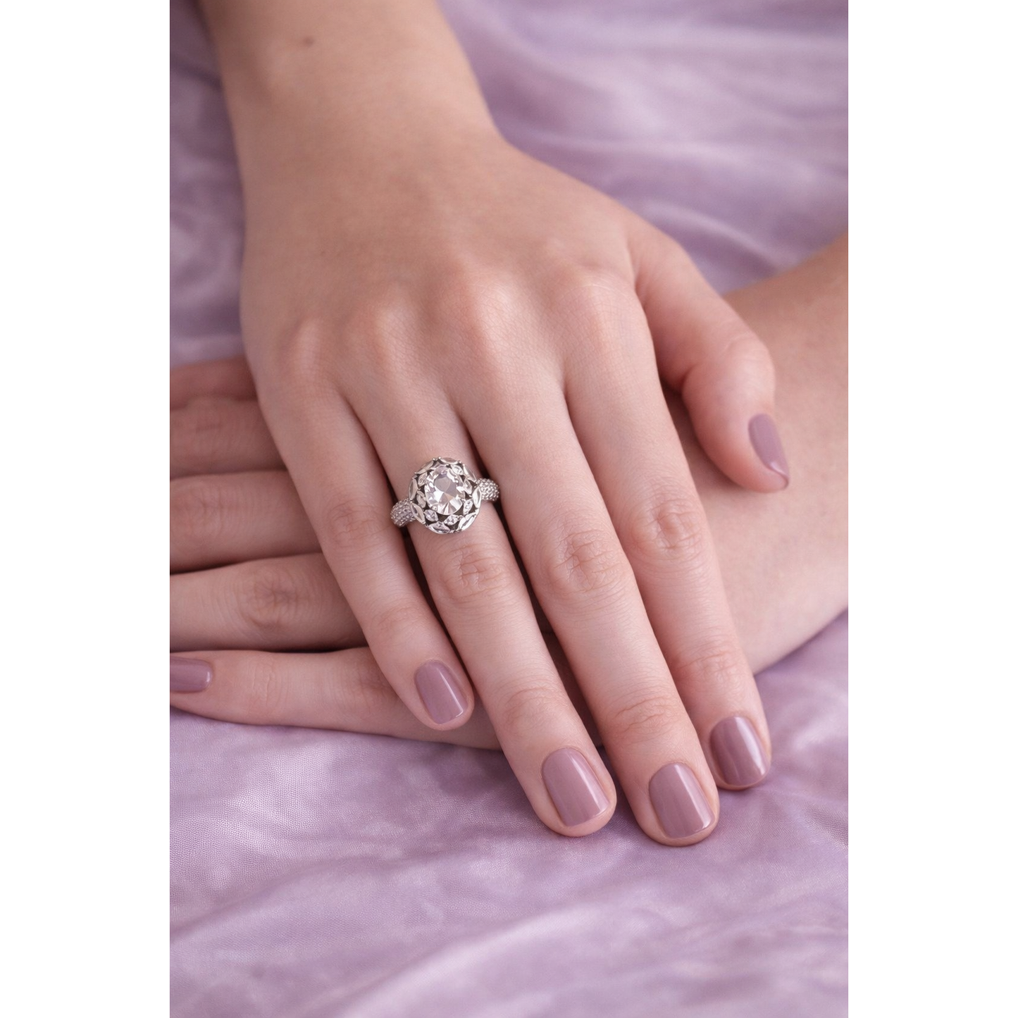 Silver round halo center stone ring worn on female hand with mauve nails against lilac silk fabric