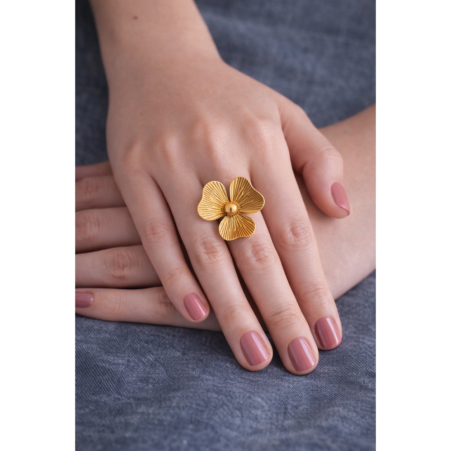 Gold flower statement ring worn on feminine hand with pink nail polish on grey denim fabric background