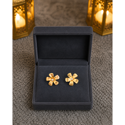 Gold floral stud earrings placed flat in dark grey velvet jewelry box on textured surface with soft lantern lighting