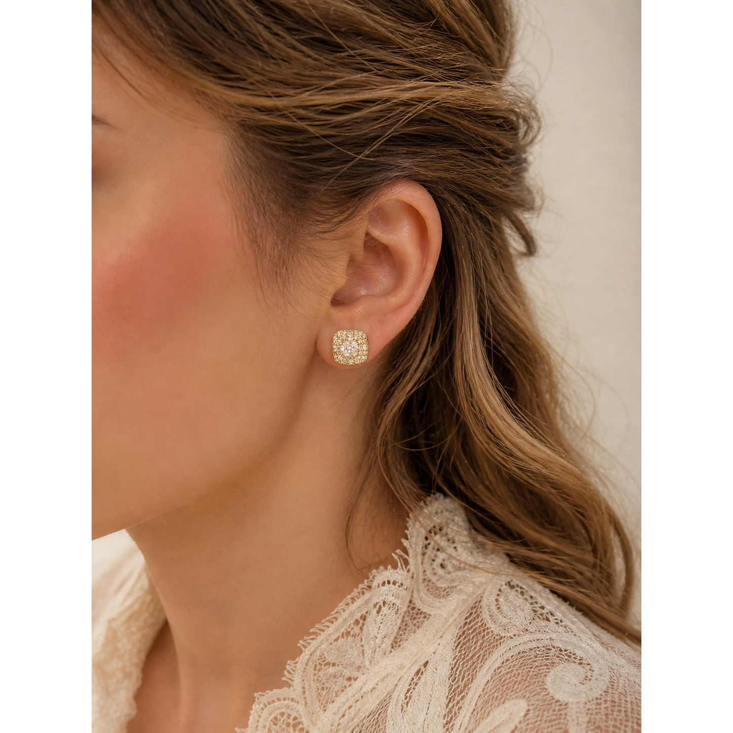 Gold cushion halo stud earrings worn on ear with light brown wavy hair and soft makeup, close-up product view