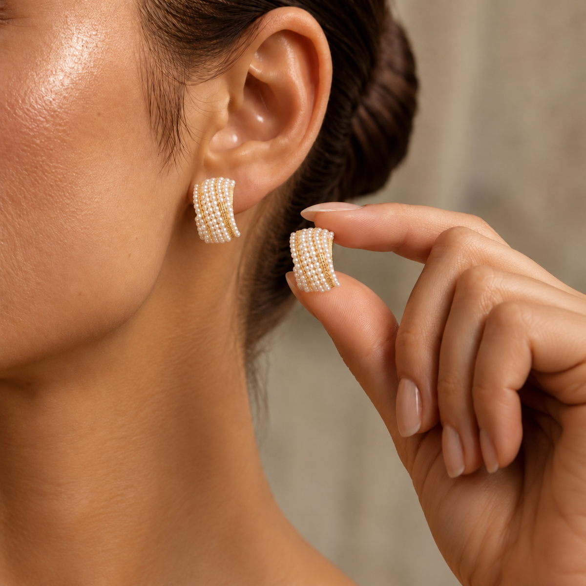 Close-up of model wearing Pearl Ripple Stud Earring while holding the matching earring, showing the curved multi-row pearl design and gold-tone accents.