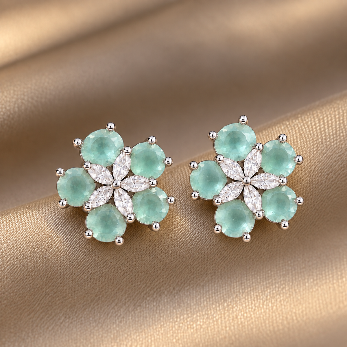 Pair of Fivefold Bloom Studs featuring five faceted  aqua blue stones surrounding a clear marquise-cut crystal flower in a polished silver-tone setting, displayed on beige satin.