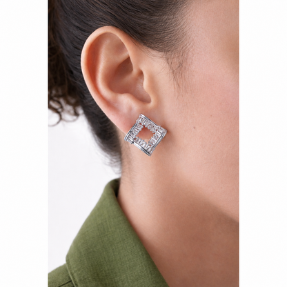 A close-up of a person's ear wearing a silver geometric square stud earring with a textured, openwork design.