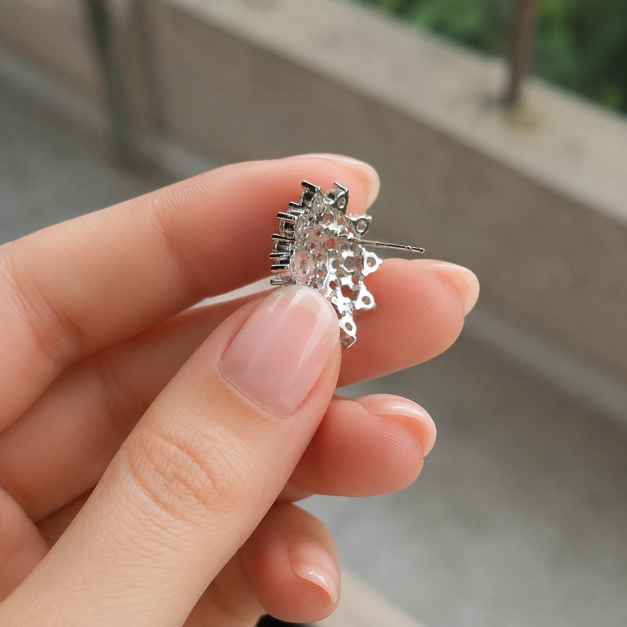 A close-up of a hand holding a small silver-toned earring with a textured, flower-like cluster setting and a clear central stone, against a blurred outdoor background.