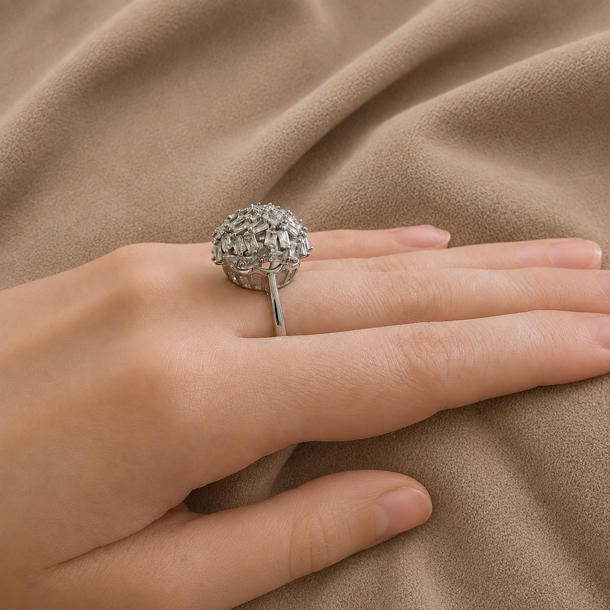 A silver-toned ring with a textured, flower-like spherical top is worn on a hand, set against a draped beige fabric background.