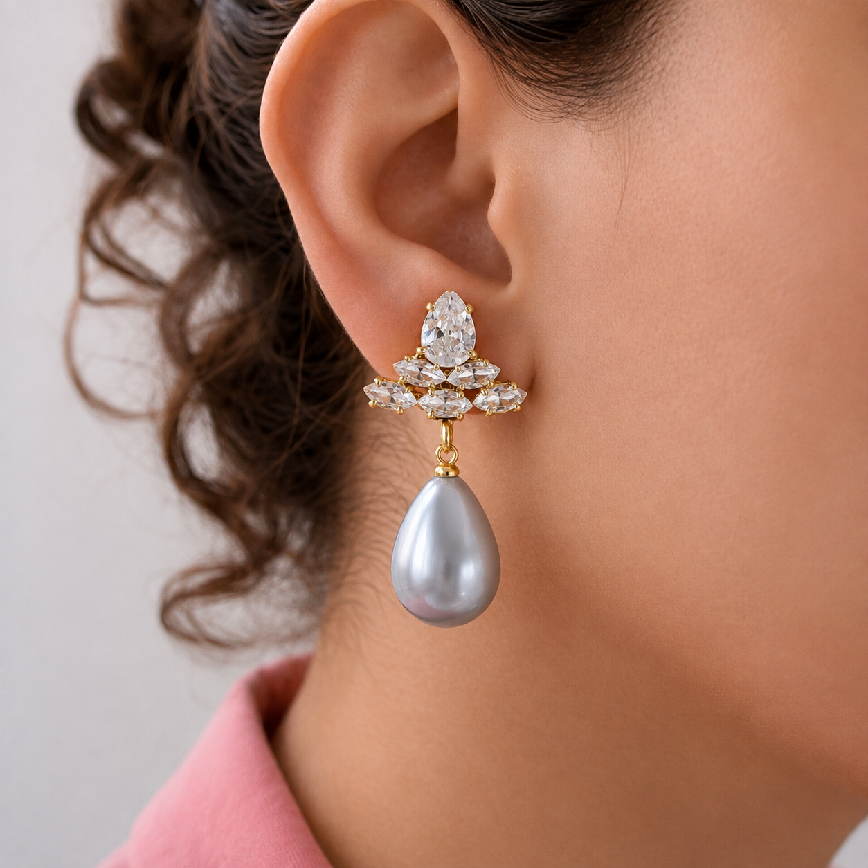 A close-up of a woman's ear wearing a gold and crystal drop earring with a teardrop-shaped gray pearl.