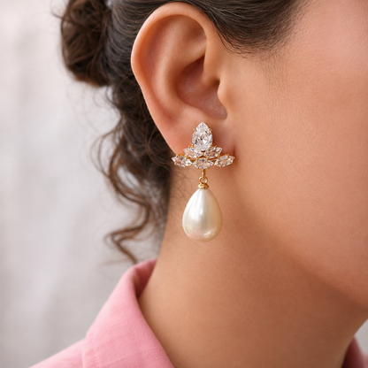 A close-up of a model's ear wearing a gold and crystal drop earring with a teardrop-shaped pearl pendant, paired with a pink collared shirt against a soft neutral background.