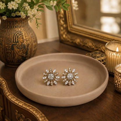 A pair of gold-toned starburst stud earrings with clear gemstones, displayed on a round beige ceramic tray on a wooden table with decorative props including a gold vase, candle, and ornate mirror.