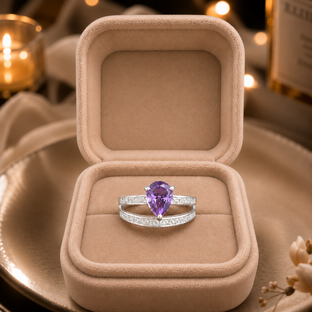 A silver ring with a pear-cut purple gemstone and pavé-set stones, displayed inside an open beige velvet ring box on a plate with warm bokeh background lights.