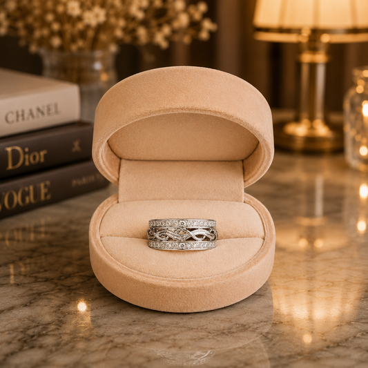A silver infinity-style ring set with small clear stones, displayed in an open beige velvet ring box on a polished marble surface with softly blurred books and a lamp in the background.