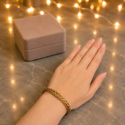 A gold braided bracelet worn on a woman's wrist, with a matching beige jewelry box in the background and warm string lights glowing on a gray marble surface.