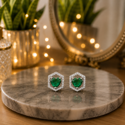 Silver hexagon stud earrings with emerald green heart-shaped crystals displayed on a polished grey marble slab with a softly blurred luxury vanity background.