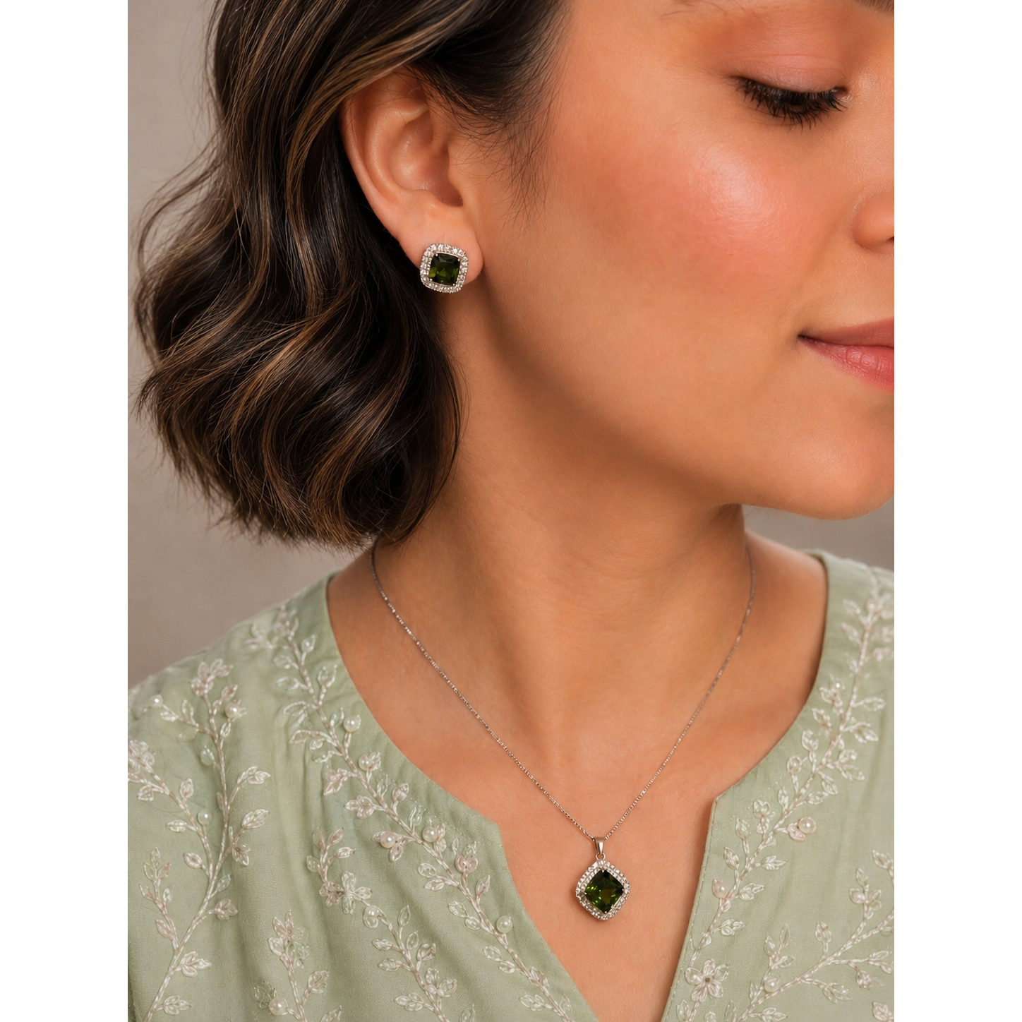 Woman wearing emerald-green crystal halo stud earrings and matching pendant necklace in a silver-tone setting with a soft pastel embroidered outfit.