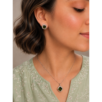 Woman wearing emerald-green crystal halo stud earrings and matching pendant necklace in a silver-tone setting with a soft pastel embroidered outfit.
