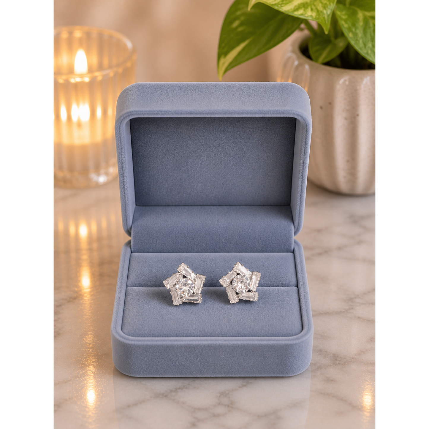 Silver baguette cluster stud earrings displayed in smoky blue velvet jewelry box on marble surface with candle and plant background
