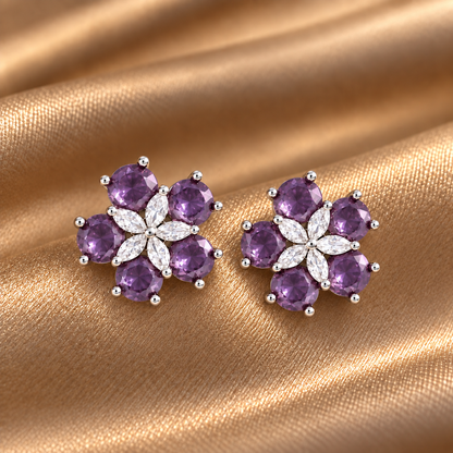 Pair of Fivefold Bloom Studs featuring five faceted  amethyst purple stones surrounding a clear marquise-cut crystal flower in a polished silver-tone setting, displayed on beige satin.