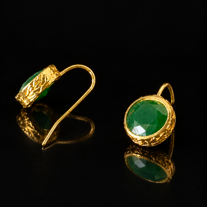 Verdant Relic Drop earrings from The Legacy Collection with round faceted emerald-green stones, textured gold-tone frames and long hook fastenings on a black reflective surface