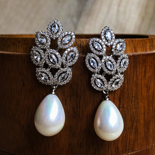 Two ornate silver drop earrings, each featuring a cluster of marquise and round clear stones above a dangling white teardrop pearl, displayed against a wooden background.