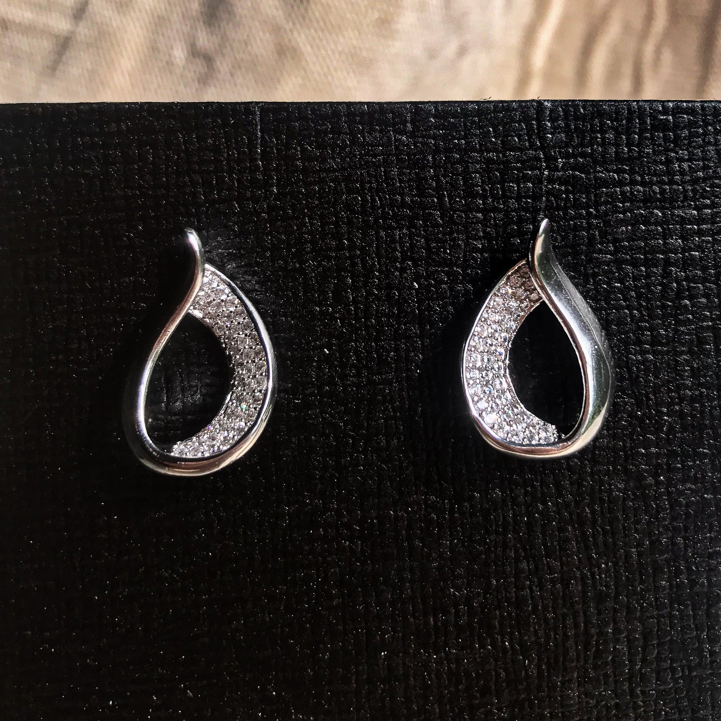 Two teardrop-shaped stud earrings, one silver and one gold, each with one side covered in sparkling clear stones, displayed on a black textured surface.