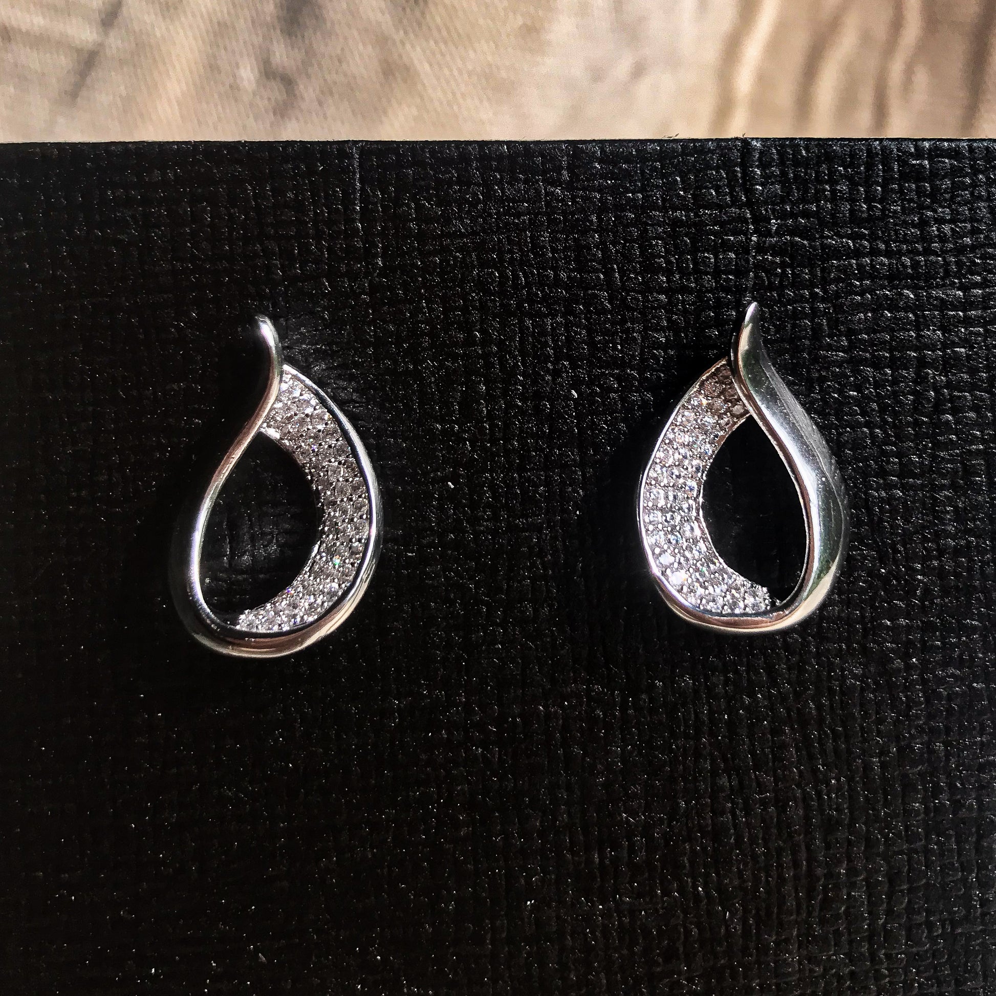 Two teardrop-shaped stud earrings, one silver and one gold, each with one side covered in sparkling clear stones, displayed on a black textured surface.