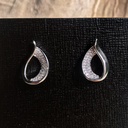 Two teardrop-shaped stud earrings, one silver and one gold, each with one side covered in sparkling clear stones, displayed on a black textured surface.