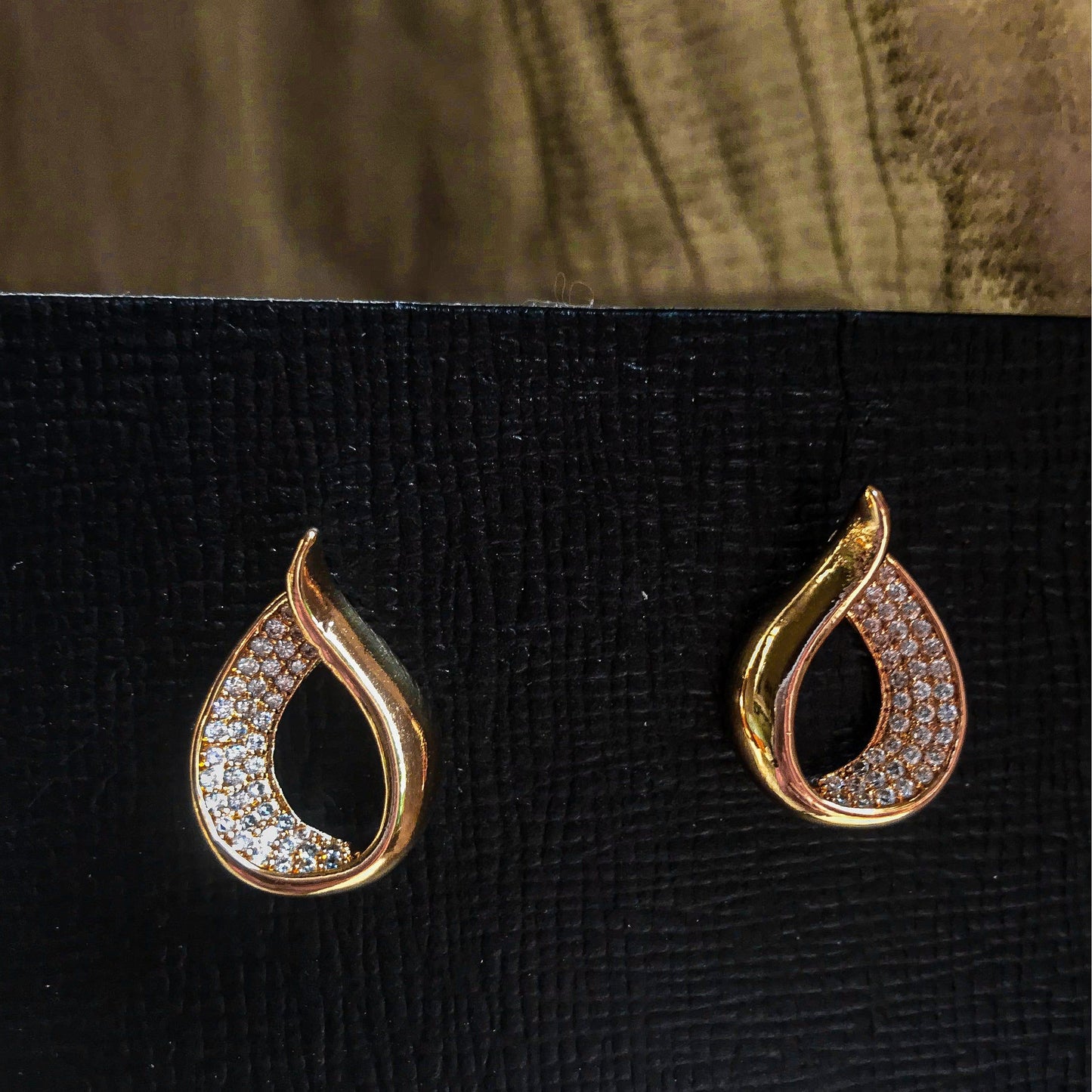 Two teardrop-shaped stud earrings, one silver and one gold, each with one side covered in sparkling clear stones, displayed on a black textured surface.