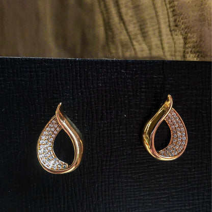 Two teardrop-shaped stud earrings, one silver and one gold, each with one side covered in sparkling clear stones, displayed on a black textured surface.