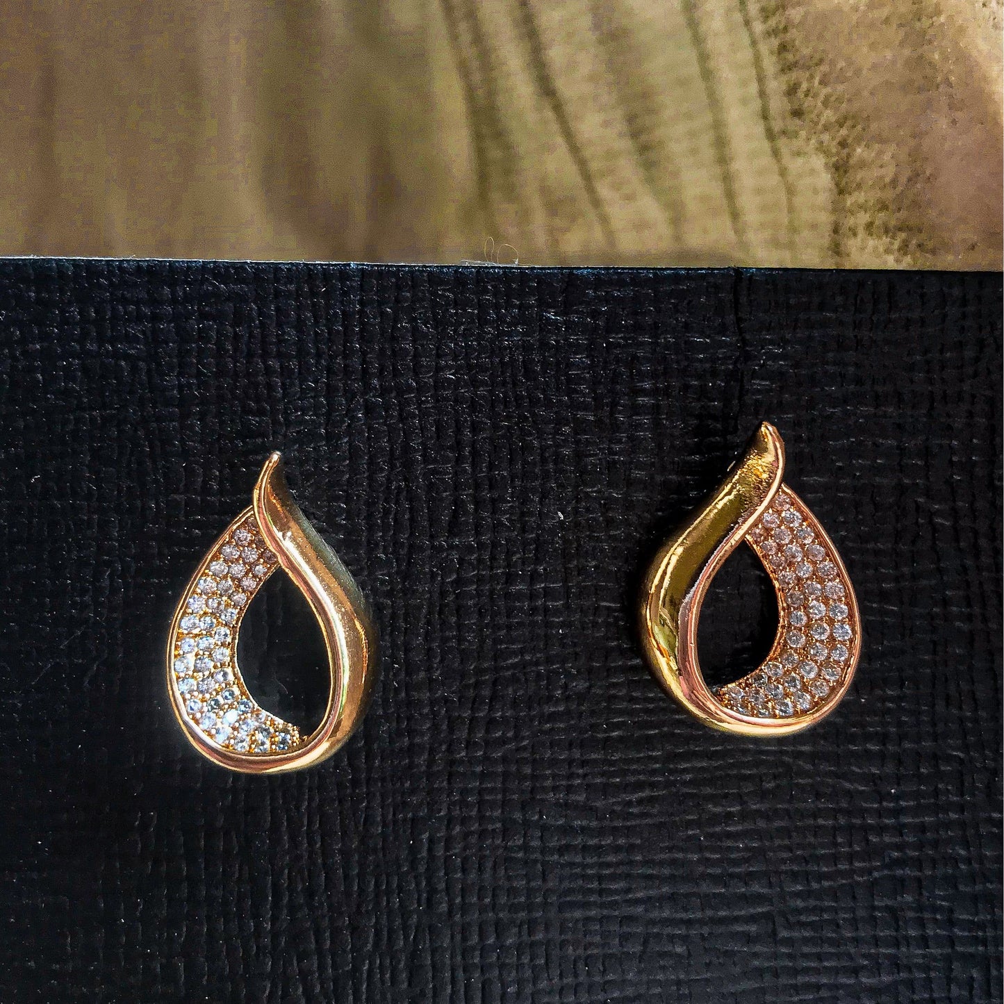Two teardrop-shaped stud earrings, one silver and one gold, each with one side covered in sparkling clear stones, displayed on a black textured surface.