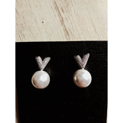 Two silver drop earrings, each featuring a white pearl hanging below a sparkling silver V-shape made of small clear stones, displayed on a textured black surface.