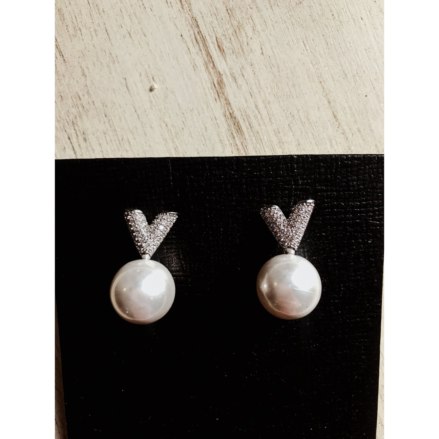 Two silver drop earrings, each featuring a white pearl hanging below a sparkling silver V-shape made of small clear stones, displayed on a textured black surface.