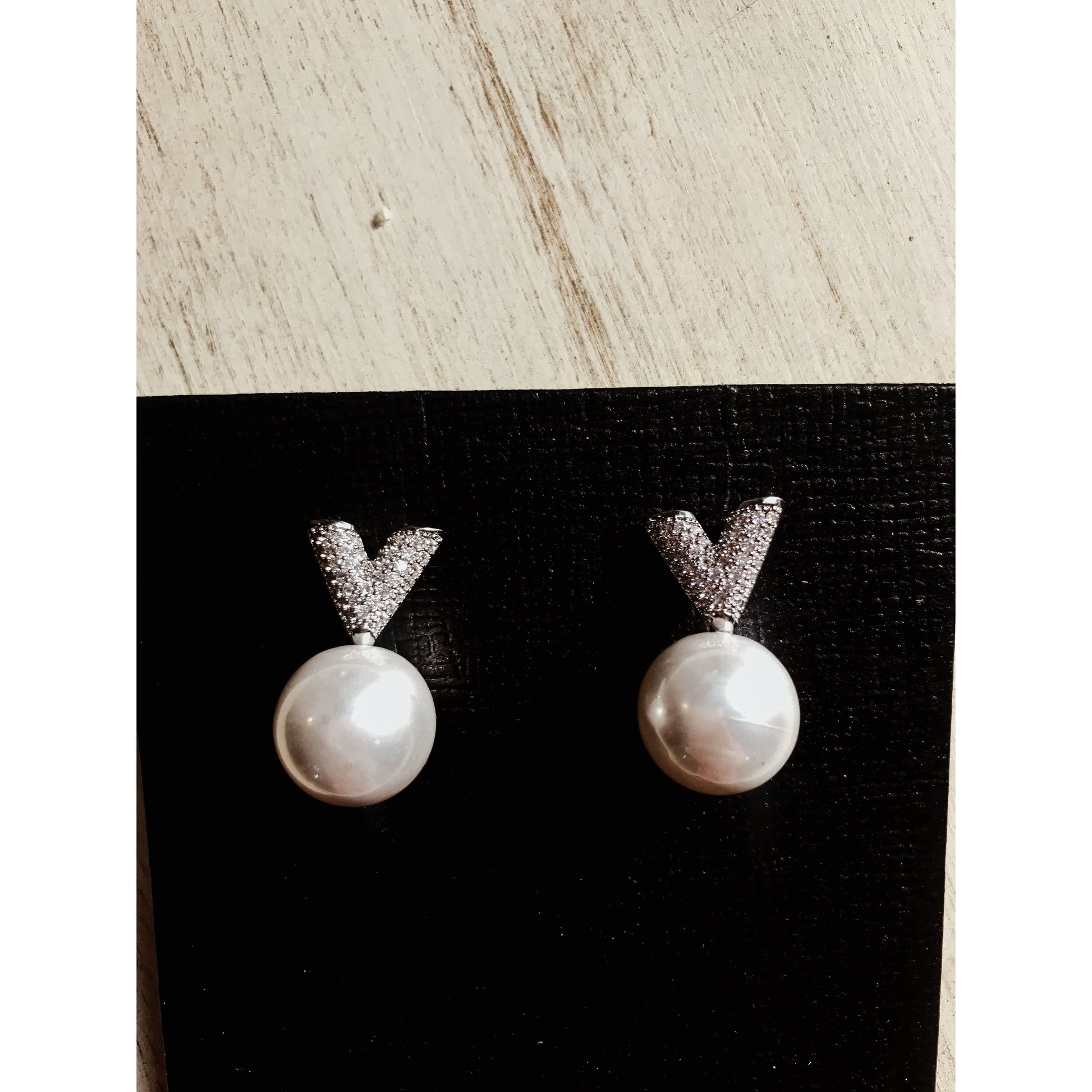 Two silver drop earrings, each featuring a white pearl hanging below a sparkling silver V-shape made of small clear stones, displayed on a textured black surface.