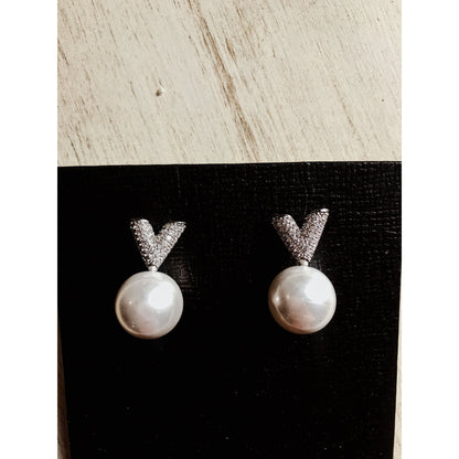 Two silver drop earrings, each featuring a white pearl hanging below a sparkling silver V-shape made of small clear stones, displayed on a textured black surface.