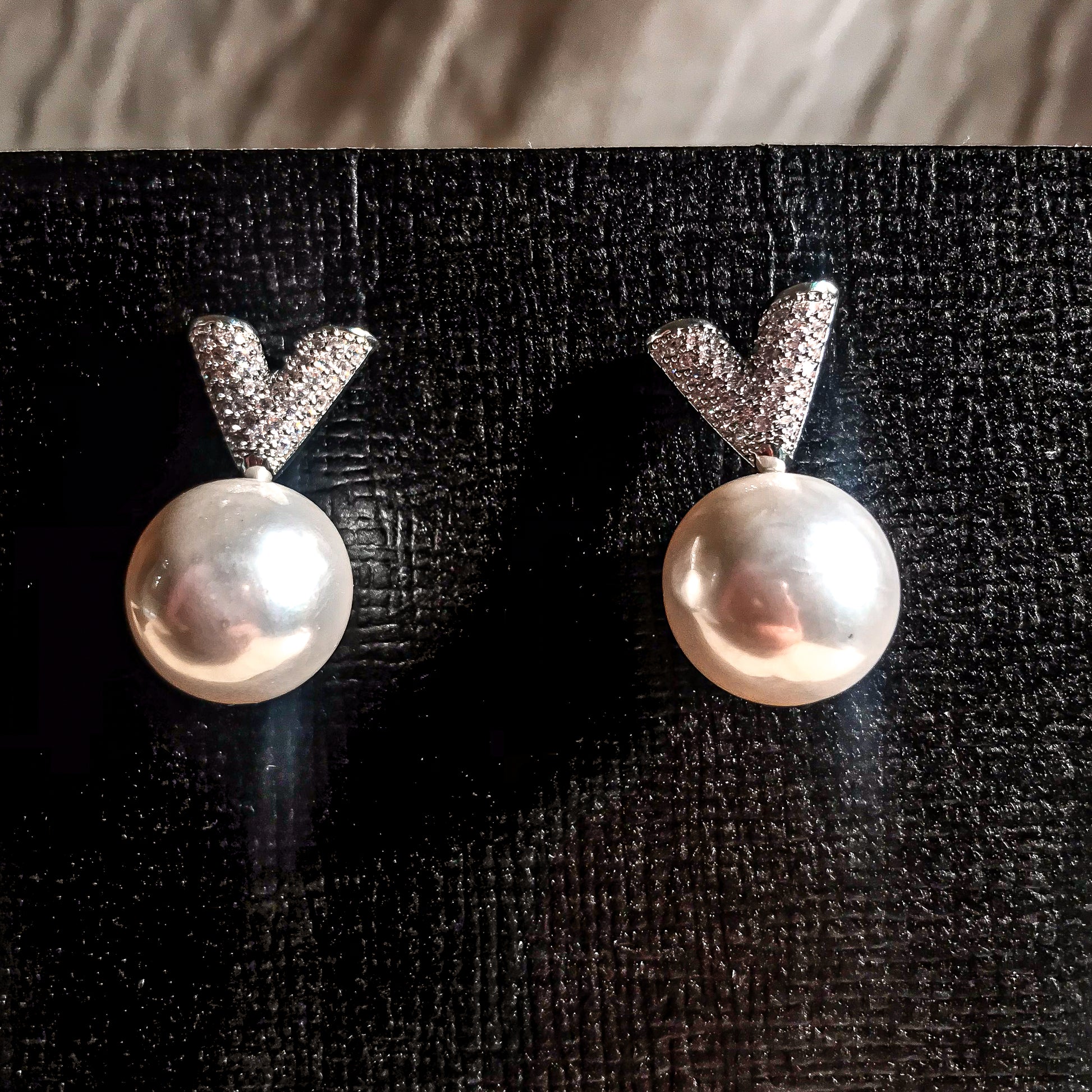 Two silver drop earrings, each featuring a white pearl hanging below a sparkling silver V-shape made of small clear stones, displayed on a textured black surface.
