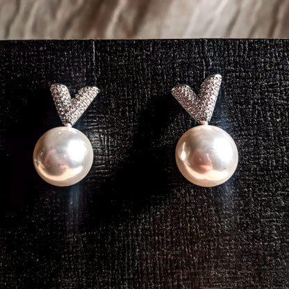 Two silver drop earrings, each featuring a white pearl hanging below a sparkling silver V-shape made of small clear stones, displayed on a textured black surface.