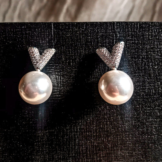 Two silver drop earrings, each featuring a white pearl hanging below a sparkling silver V-shape made of small clear stones, displayed on a textured black surface.
