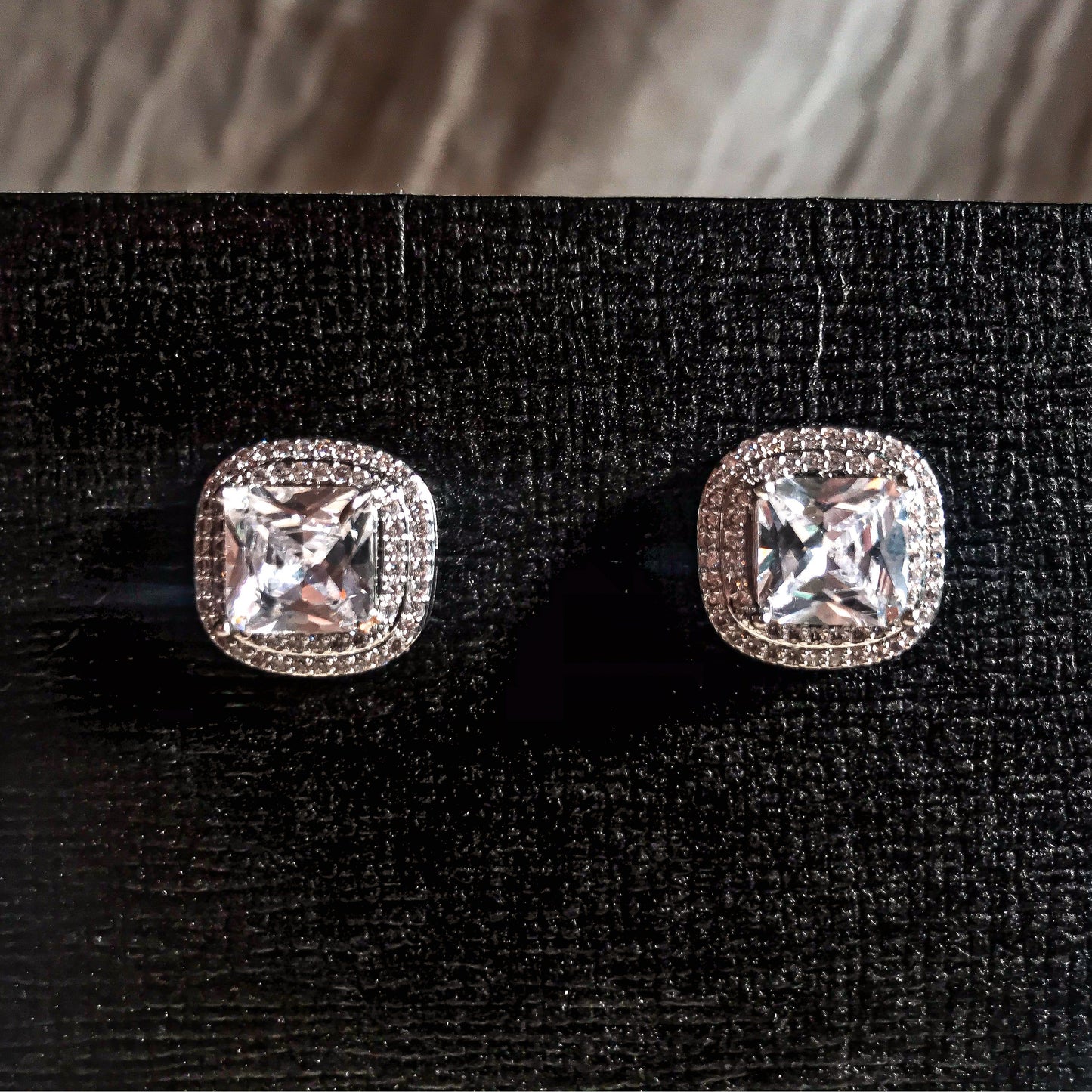Two silver stud earrings, each featuring a large, square cushion-cut clear stone surrounded by smaller sparkling stones, displayed on a black textured surface.