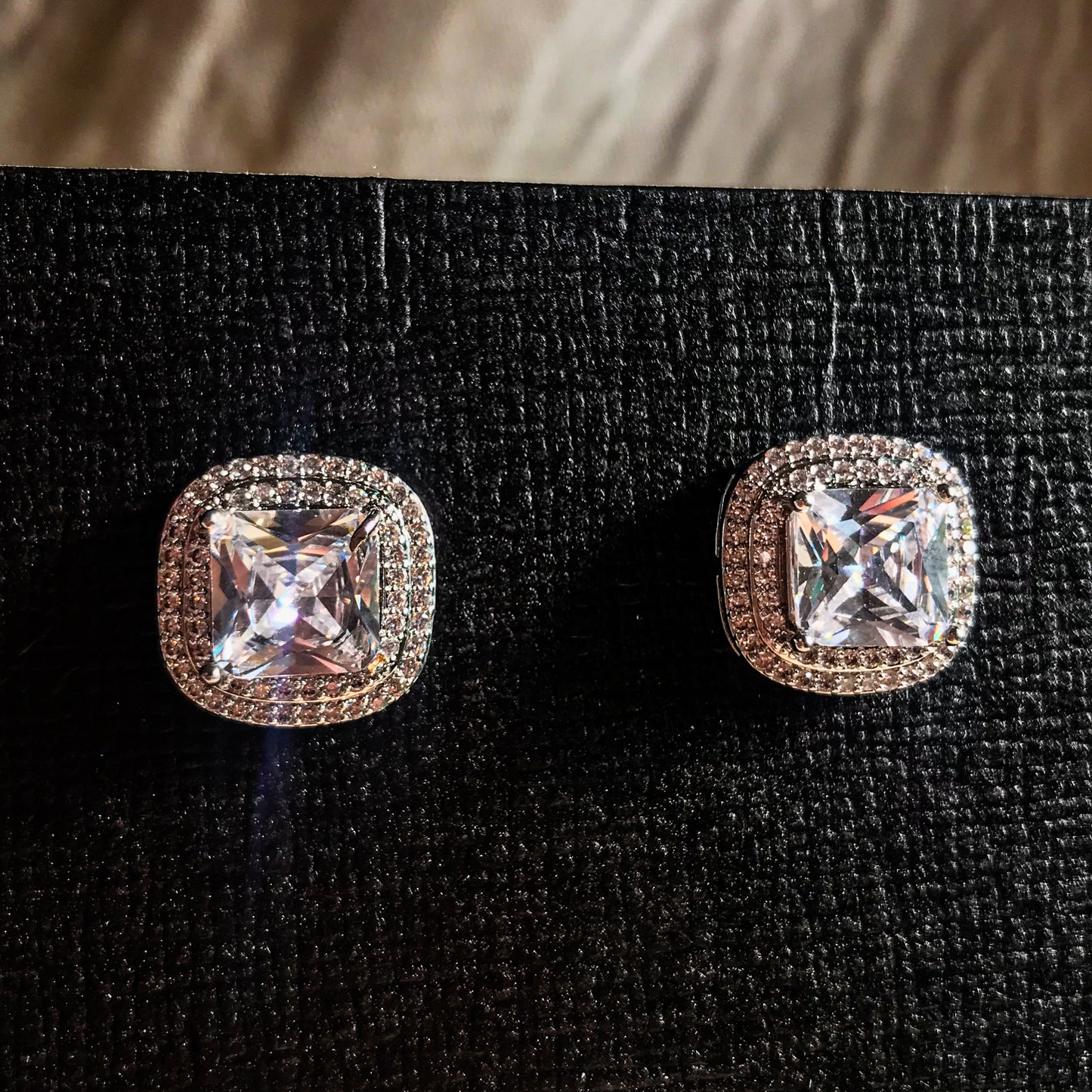 Two silver stud earrings, each featuring a large, square cushion-cut clear stone surrounded by smaller sparkling stones, displayed on a black textured surface.