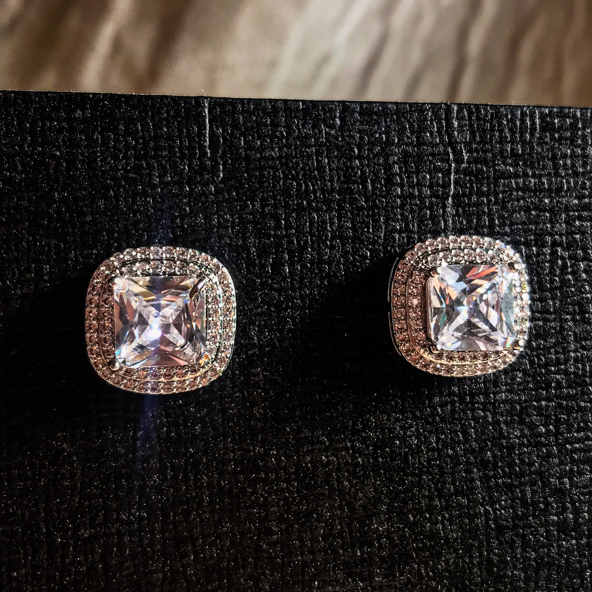 Two silver stud earrings, each featuring a large, square cushion-cut clear stone surrounded by smaller sparkling stones, displayed on a black textured surface.