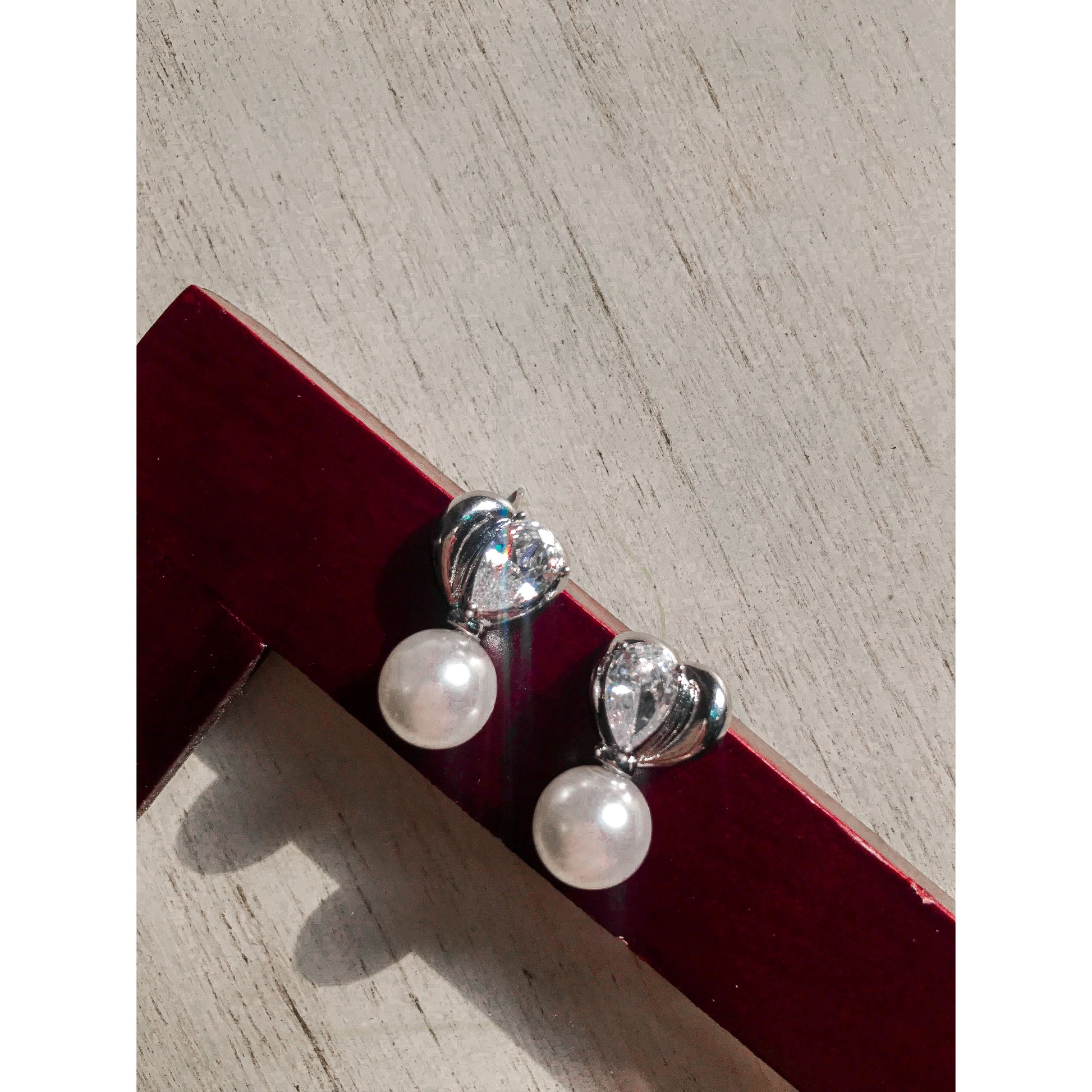 Two elegant silver drop earrings, each featuring a white pearl hanging below a silver heart with a clear, pear-shaped stone, displayed on a textured surface.